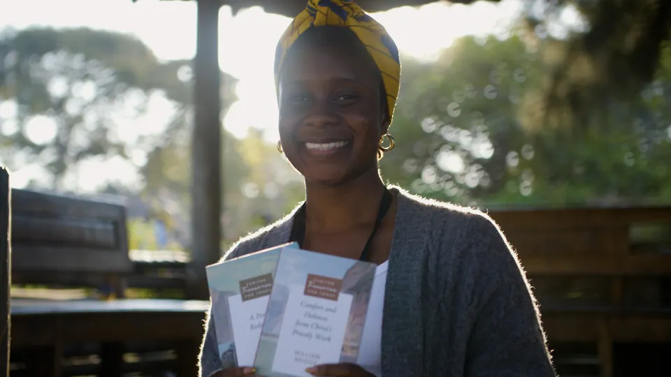 A woman in Zambia smiles in late afternoon light, holding two Puritan Treasures for Today books.