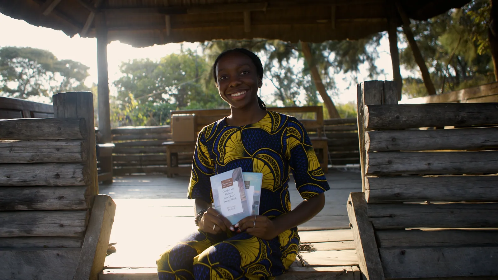 A woman in a patterned dress sits in a thatched shelter at golden hour, holding her two new books.